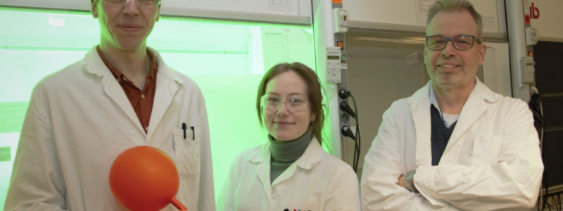 Jesper Schwarz, Aleksandra Ilic och Kenneth Wärnmark visar upp sitt röda järnkomplex inuti en vätgasballong framför en ljusreaktor. På bilden saknas Reiner Lomoth och Catherine Johnson från Uppsala universitet. Foto: Johan Joelsson